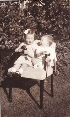Barbara and Rod Rowe as young children sitting on a garden chair, girl holding a toy, leafy bush behind, c.1940s–1950s