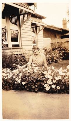 Mary Webber tending flowers in a garden in front of a weatherboard house, sepia, c.1930s–1940s