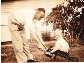 Robertson/Webber family man bending down to a small child sitting in a toy cart or wagon in a suburban garden, sepia, c.1940s–1950s