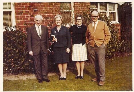 Colour photograph: four people standing in front of a brick house garden, c.1960s–1970s — two men and two women in casual dress on a suburban lawn