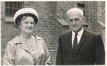 Robertson/Webber family — man and woman standing together outside a brick building, woman in floral dress and white hat, man in dark suit, c.1960s