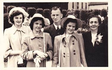 Five people outdoors: two young women in light coats (left), young man in dark suit (centre-back), young woman in hat and dark coat (centre-right), older woman in dark coat with corsage (far right) — c.1940s–1950s