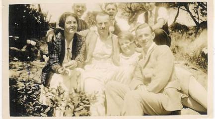 Six people seated and reclining on rocks or a hillside — two women at left (one in patterned jacket), man in centre, woman in white, man at right — trees and bush behind — c.1920s–1930s