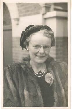 Mary Webber — outdoor/street portrait, middle-aged woman in dark coat with fur-trimmed collar and small decorative hat, smiling broadly