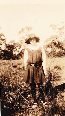 Woman in sleeveless dress and wide-brimmed hat standing in a field with gum trees behind, c.1920s — outdoor portrait in Australian bush setting