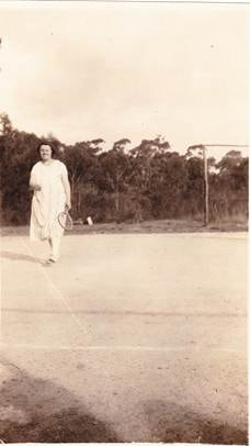 Robertson/Webber family woman in white dress on a tennis court, racquet in hand, bush behind, c.1920s–1930s